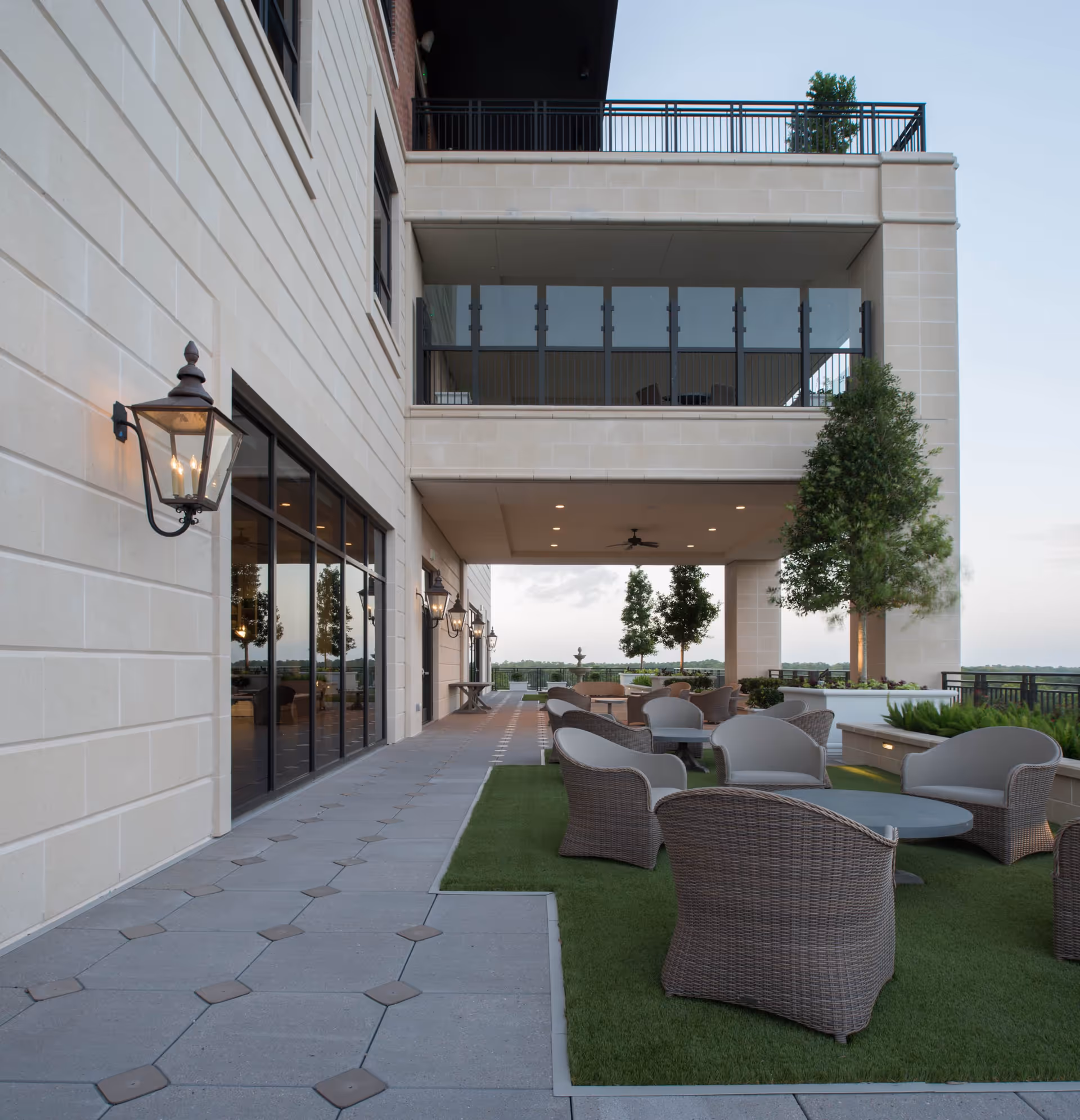 Outdoor patio area at The Tradition–Woodway featuring wicker chairs and tables arranged on artificial grass and tiled flooring, with large windows and wall-mounted lantern lights on the building exterior, and trees and a railing overlooking a distant landscape.