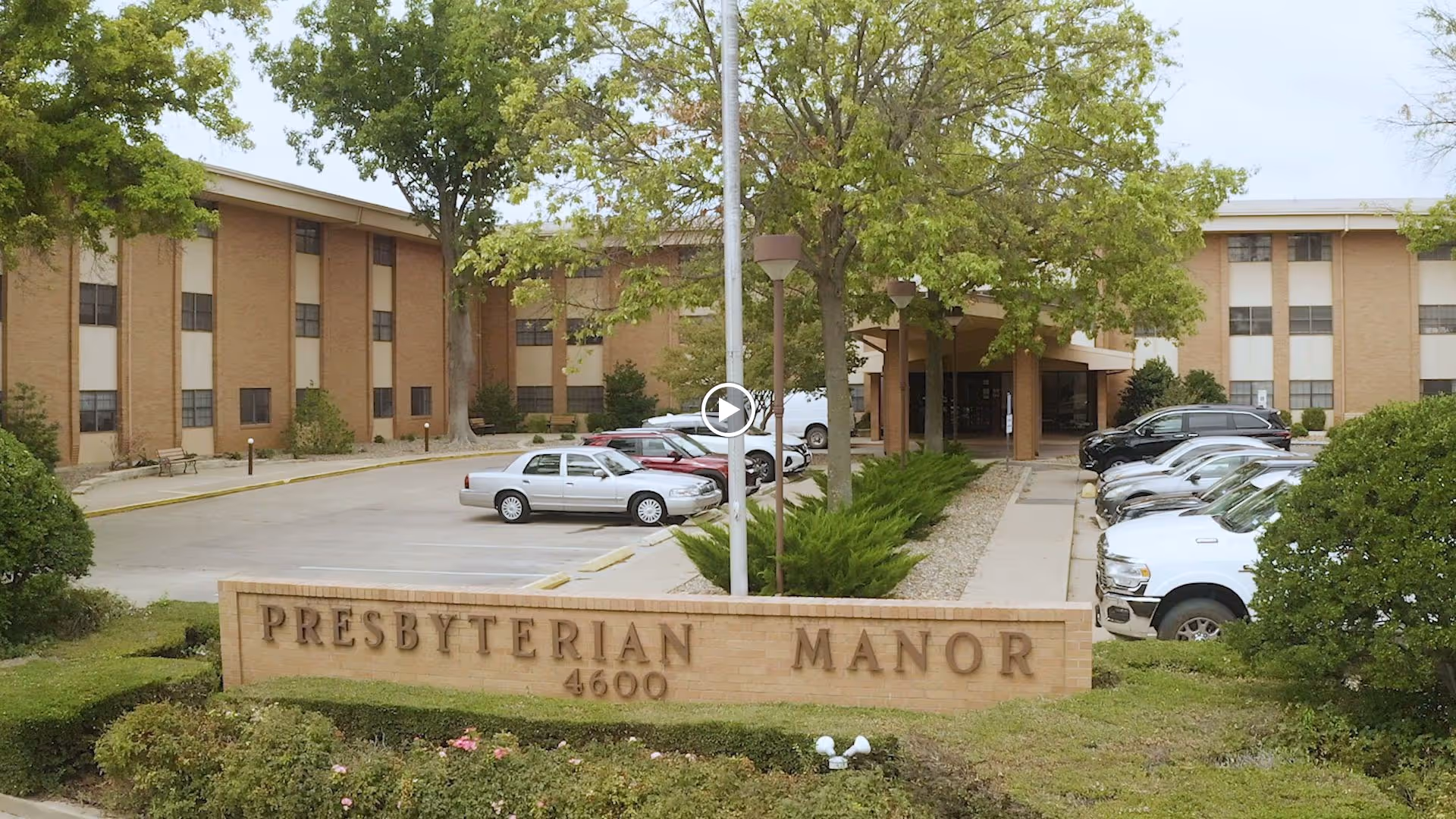 Front entrance of Presbyterian Manor with a brick sign reading "PRESBYTERIAN MANOR 4600", a driveway and parked cars in front of the three-story building.
