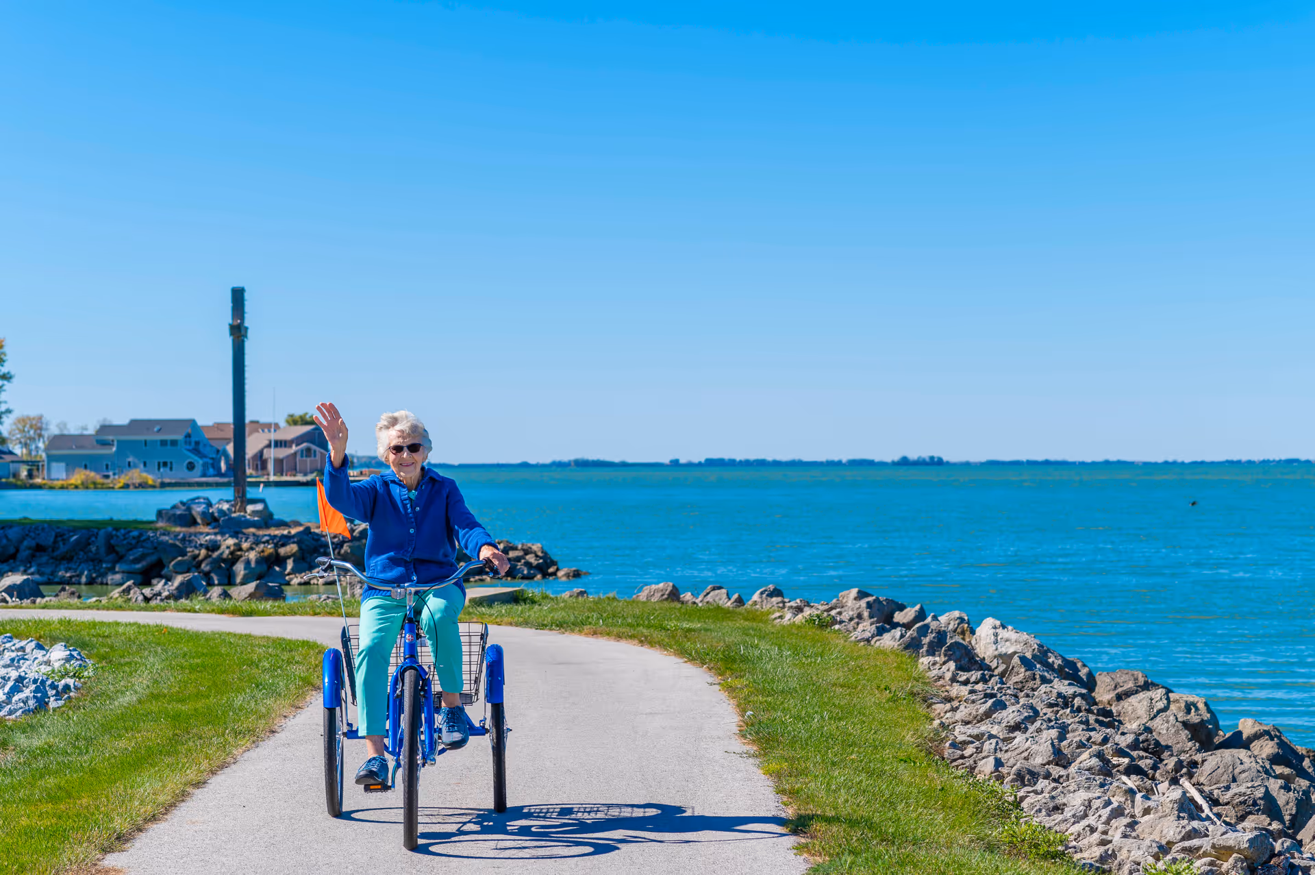 An elderly woman wearing sunglasses and a blue jacket rides a blue three-wheeled bicycle on a paved path along a rocky shoreline with a large body of water and houses in the background. She is smiling and waving with her left hand.