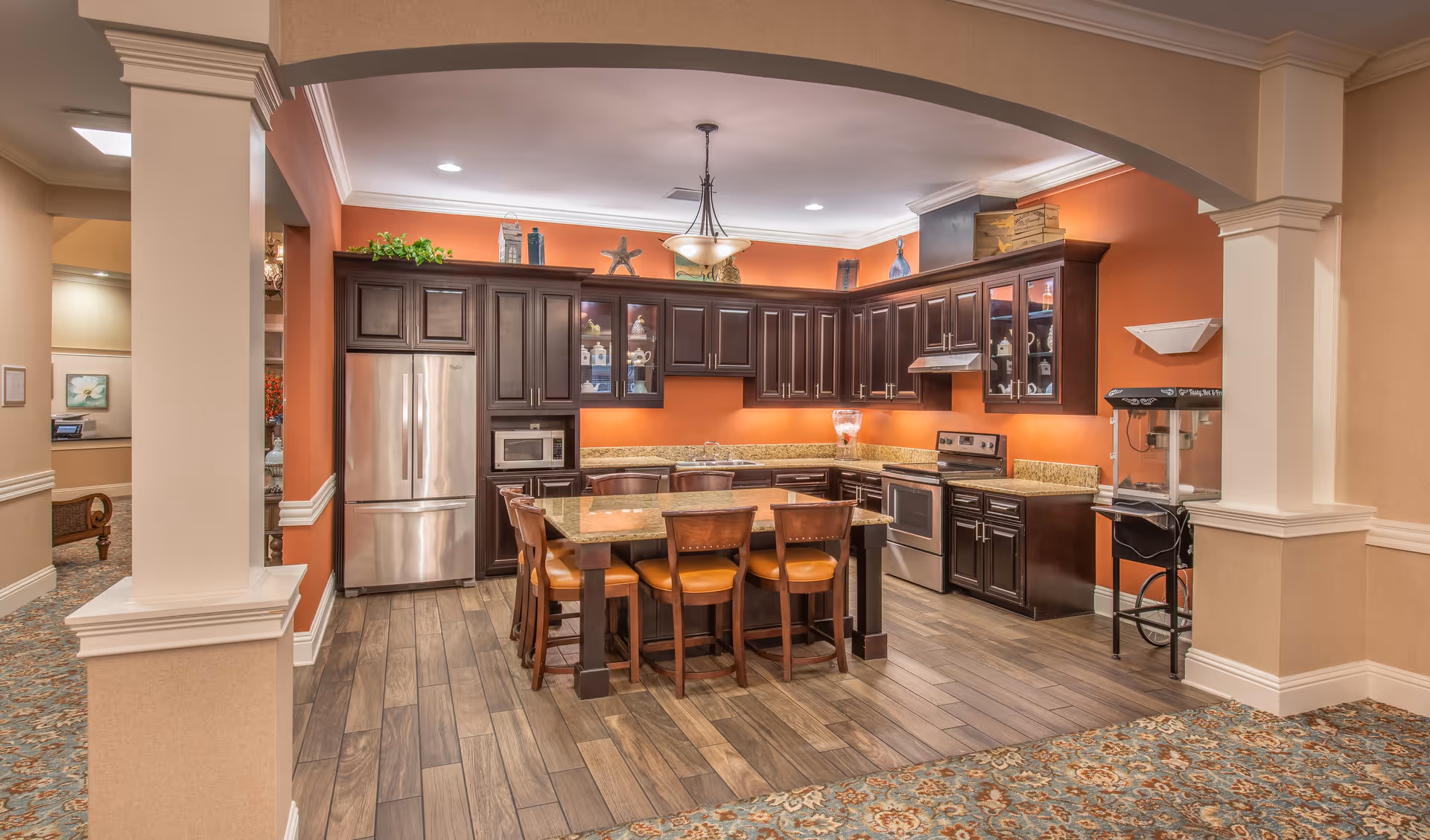 A spacious kitchen area with dark wood cabinets, stainless steel refrigerator, microwave, stove, and a central island with six wooden chairs. The walls are painted orange, and the floor has wood-like tiles. There is a popcorn machine on the right side and decorative items on top of the cabinets. The kitchen is framed by beige walls and an archway.