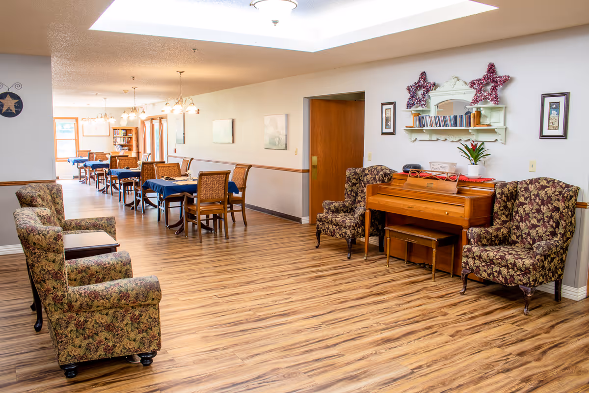 A spacious senior living facility common area with wooden flooring, floral upholstered armchairs, a wooden piano with a small bench, and several dining tables covered with blue tablecloths. The walls are decorated with framed pictures and a shelf holding books and decorative stars. The room is well-lit with ceiling lights and natural light from windows in the background.