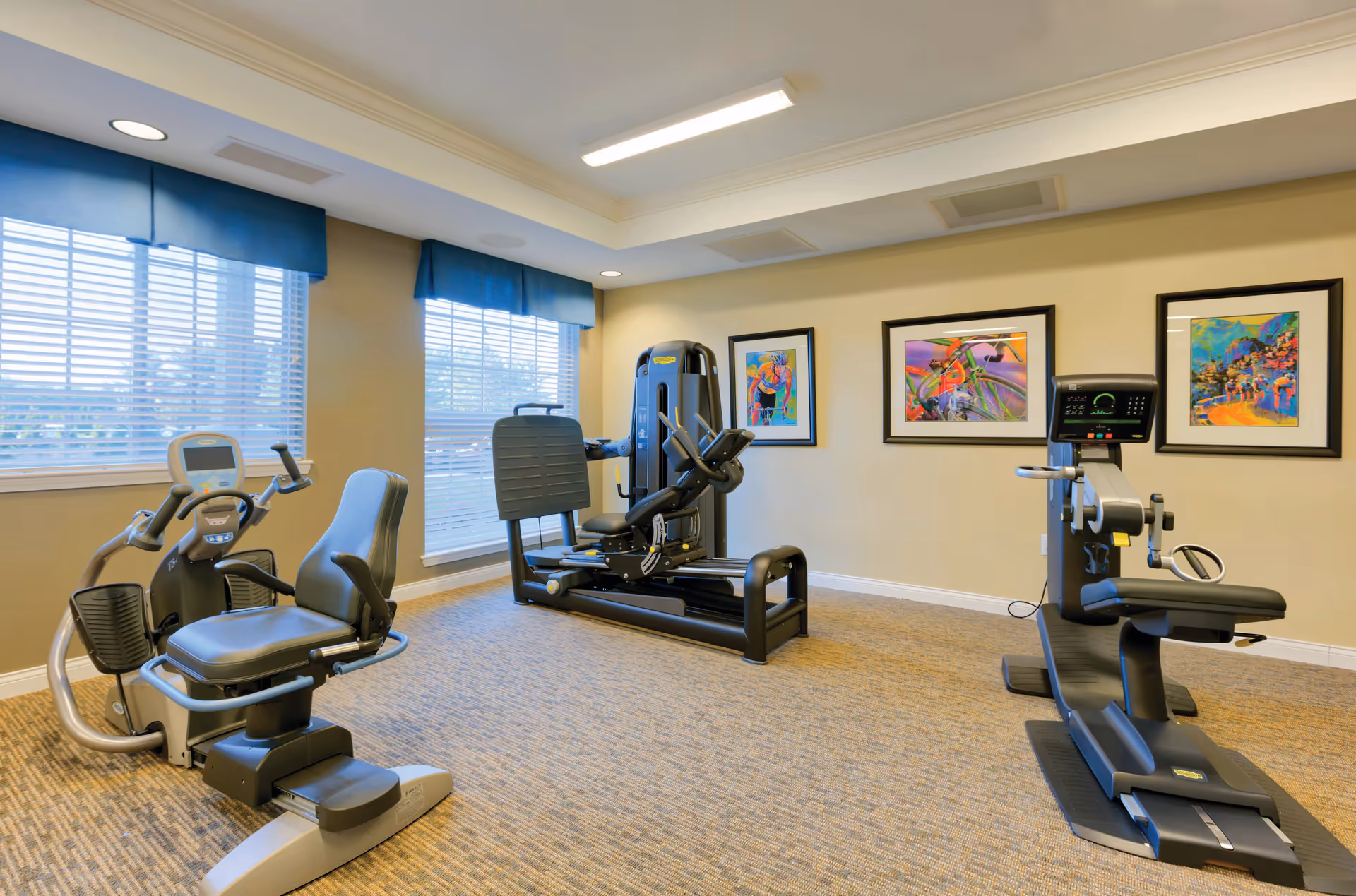 Small fitness room with exercise machines and recumbent bikes, windows with blinds, and framed artwork on the wall.