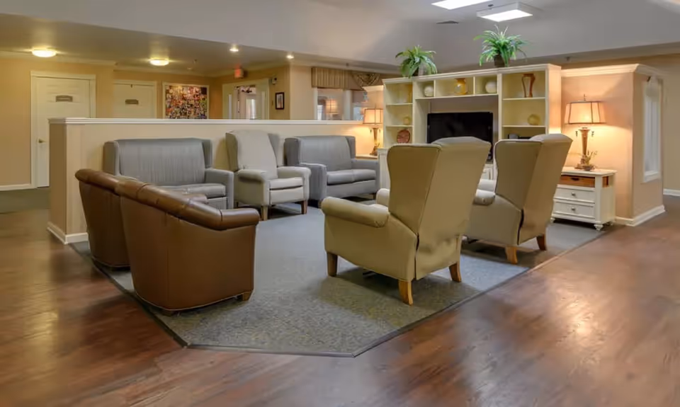 A cozy senior living facility common area with multiple armchairs and sofas arranged around a carpeted section. The room features wooden flooring, a built-in shelving unit with decorative items, and warm lighting from table lamps.