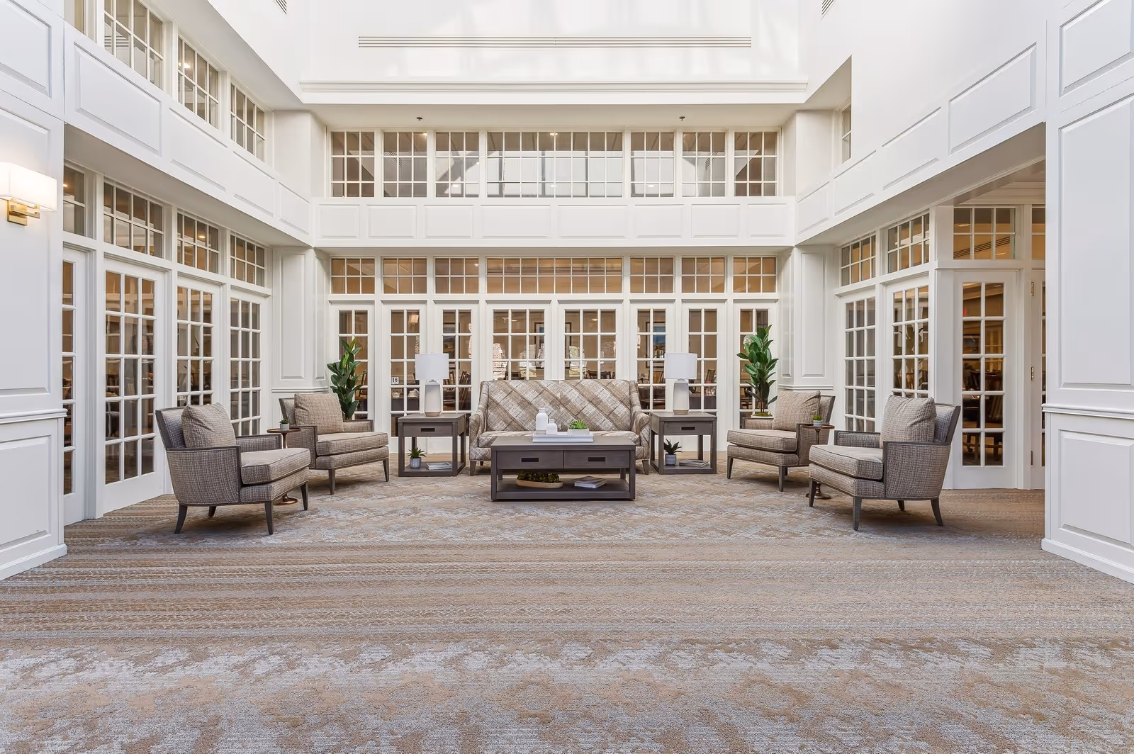 Bright, elegant communal lounge with upholstered chairs, sofas, coffee tables, and large windows under a skylight.