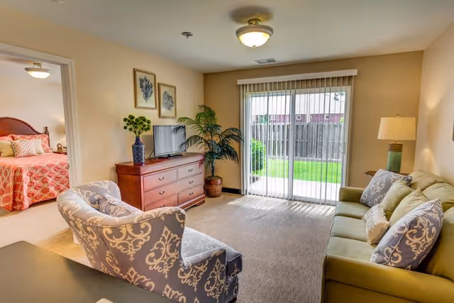 A cozy living room with a green sofa adorned with patterned pillows, a patterned armchair, a wooden dresser with a flat-screen TV, and a potted plant. Sliding glass doors with vertical blinds open to a small fenced backyard with green grass. A bedroom with a bed covered in a red patterned bedspread is visible through an open doorway.