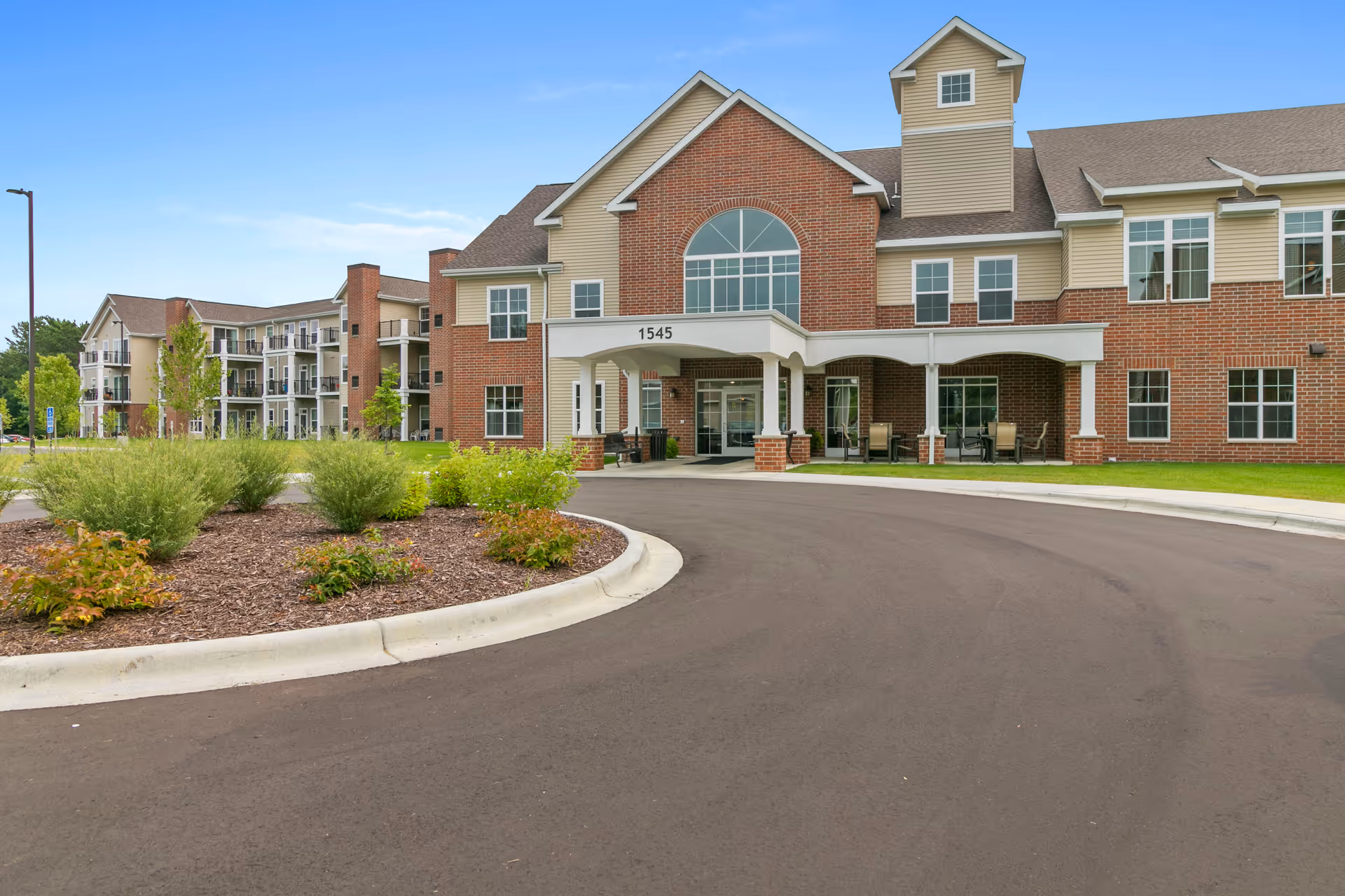 Front entrance and porte-cochère of a multi-story brick senior living building with a curved driveway and landscaped island.