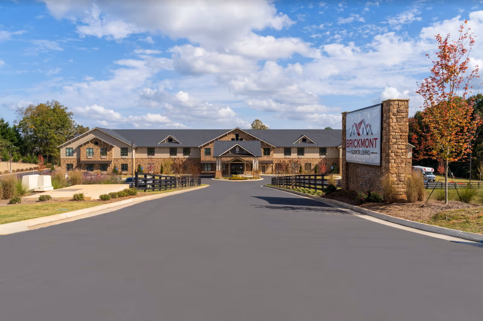 Exterior view of Brickmont at Johns Creek senior living facility with a paved driveway leading to the entrance, a stone sign displaying the facility name, and landscaping with trees and shrubs under a partly cloudy sky.