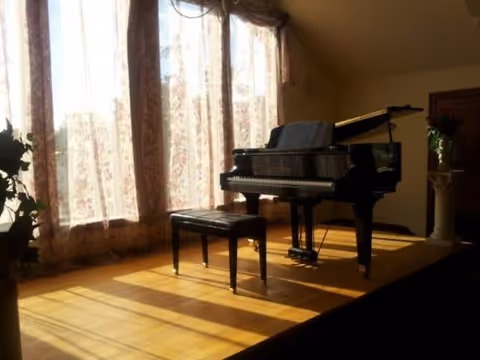 A grand piano with a matching bench is positioned on a wooden floor in front of large windows covered with sheer floral curtains, allowing sunlight to cast shadows across the floor.