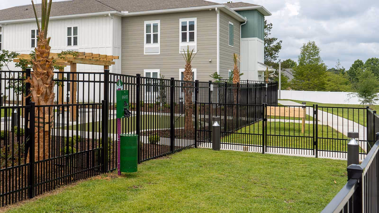 Outdoor fenced area with green grass and a black metal fence surrounding it. There are palm trees planted inside the fenced area and a green dog waste station attached to the fence. In the background, there is a two-story building with beige and white siding and multiple windows. A paved walkway and some landscaping are also visible.