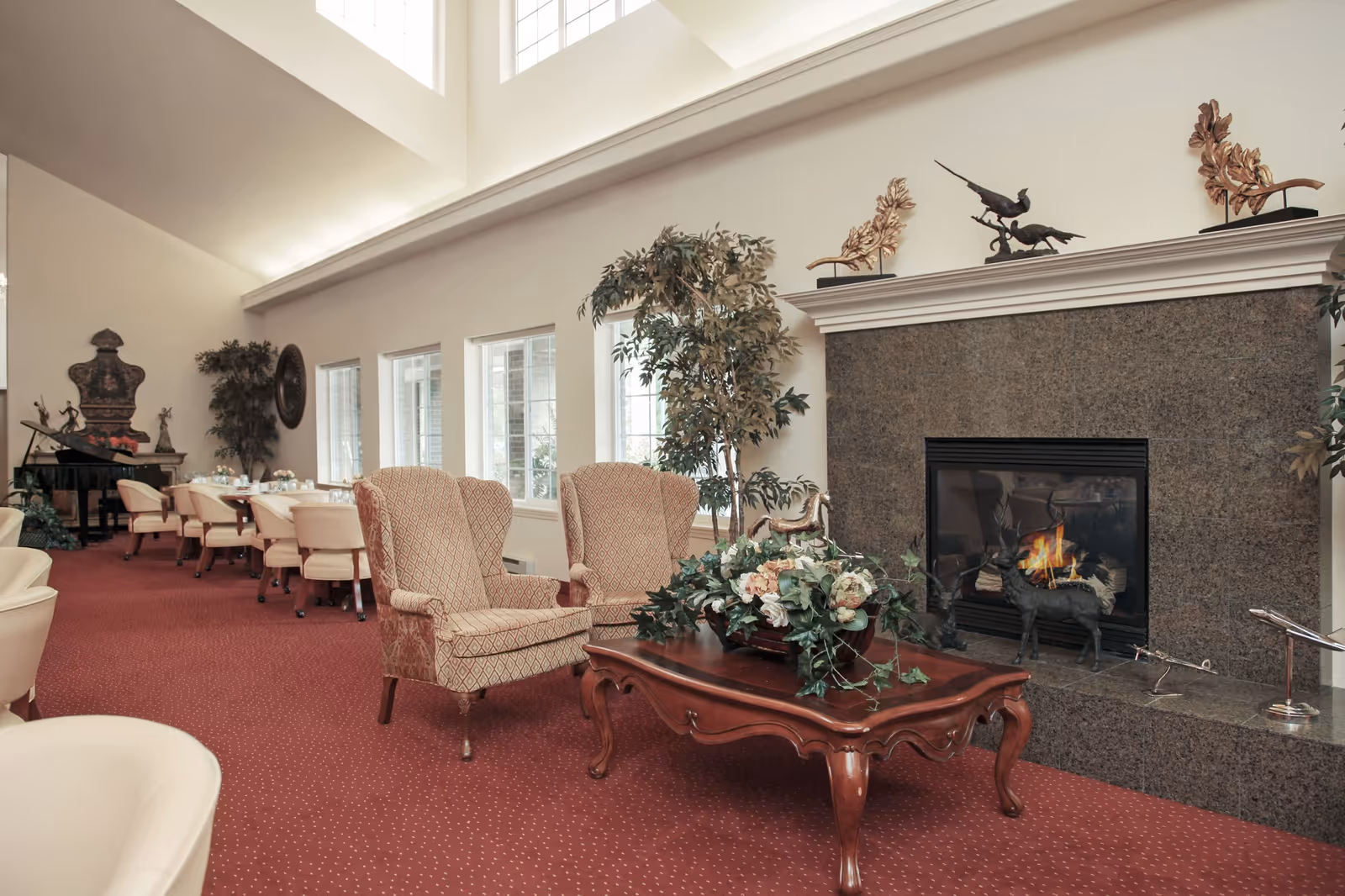 A spacious communal lounge with upholstered armchairs, a wooden coffee table, a lit fireplace and dining tables along tall windows.