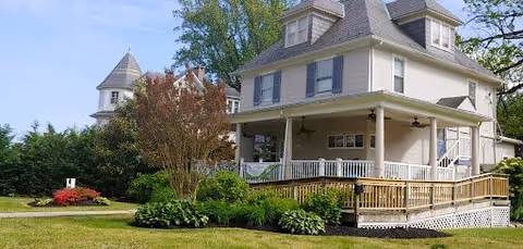 A large, light-colored two-story house with a wraparound porch and a wooden ramp leading to the entrance. The house is surrounded by green grass, bushes, and trees under a clear blue sky.