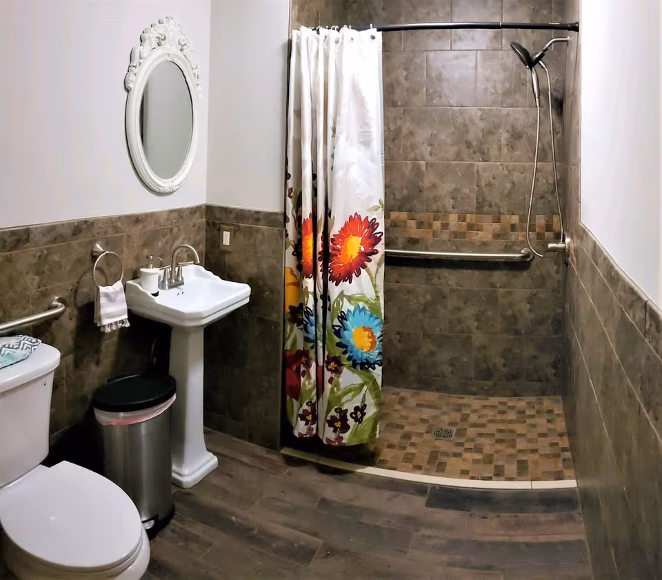 A bathroom with a white toilet, a pedestal sink with a soap dispenser, a decorative white oval mirror above the sink, a metal trash can, and a walk-in shower with brown tiled walls and floor. The shower has a grab bar and a shower curtain with colorful floral patterns.