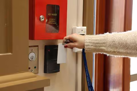 Person's arm swipes a key card near a door security panel beside a red fire cabinet.