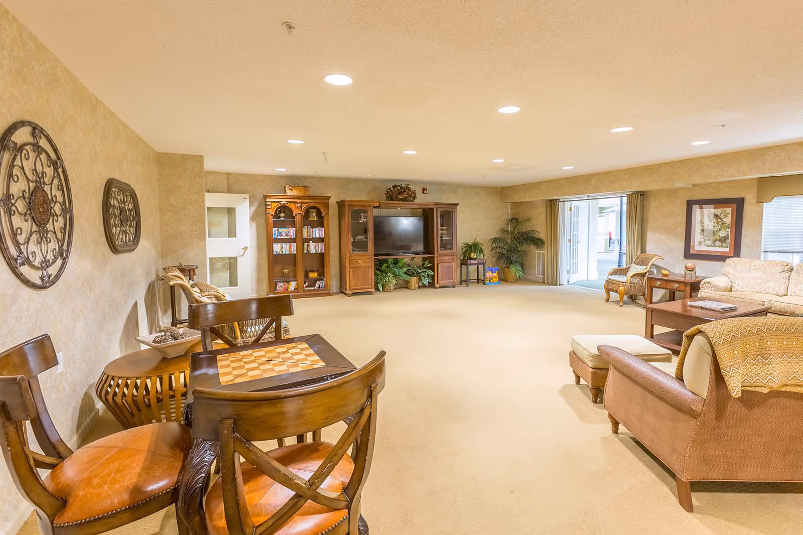 A spacious and well-lit common living area with beige carpet and walls. The room features a wooden entertainment center with a large flat-screen TV, bookshelves, and several potted plants. There are multiple seating options including a sofa, armchairs, and a small table with chairs that has a chessboard on top. Decorative wall art and framed pictures adorn the walls. Large windows and a glass door allow natural light to enter the room.
