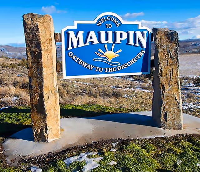 A blue and white welcome sign reading 'Welcome to Maupin, Gateway to the Deschutes' mounted between two large stone pillars, set in a natural landscape with hills and sparse vegetation under a partly cloudy sky.