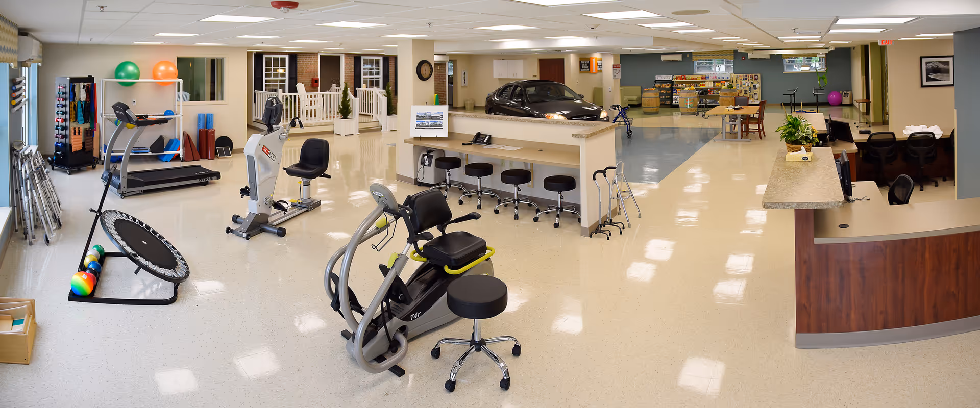 A spacious and well-lit rehabilitation or physical therapy room in a senior living facility. The room contains exercise equipment including a treadmill, stationary bikes, a mini trampoline, and various therapy balls. There are stools lined up along a counter, a desk area with chairs, and a small car used for therapy purposes. The walls are decorated with windows and a clock, and the floor is clean and shiny.
