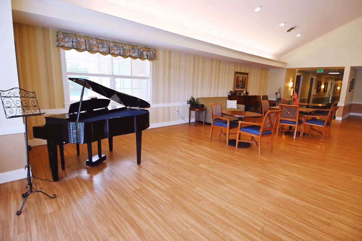 A spacious room with a black grand piano and a music stand near a large window with patterned valance. The room has wooden flooring, several round tables with wooden chairs that have blue cushions, a small couch, and a side table with a plant. The walls are decorated with striped wallpaper and a framed picture, and there is a hallway visible in the background.