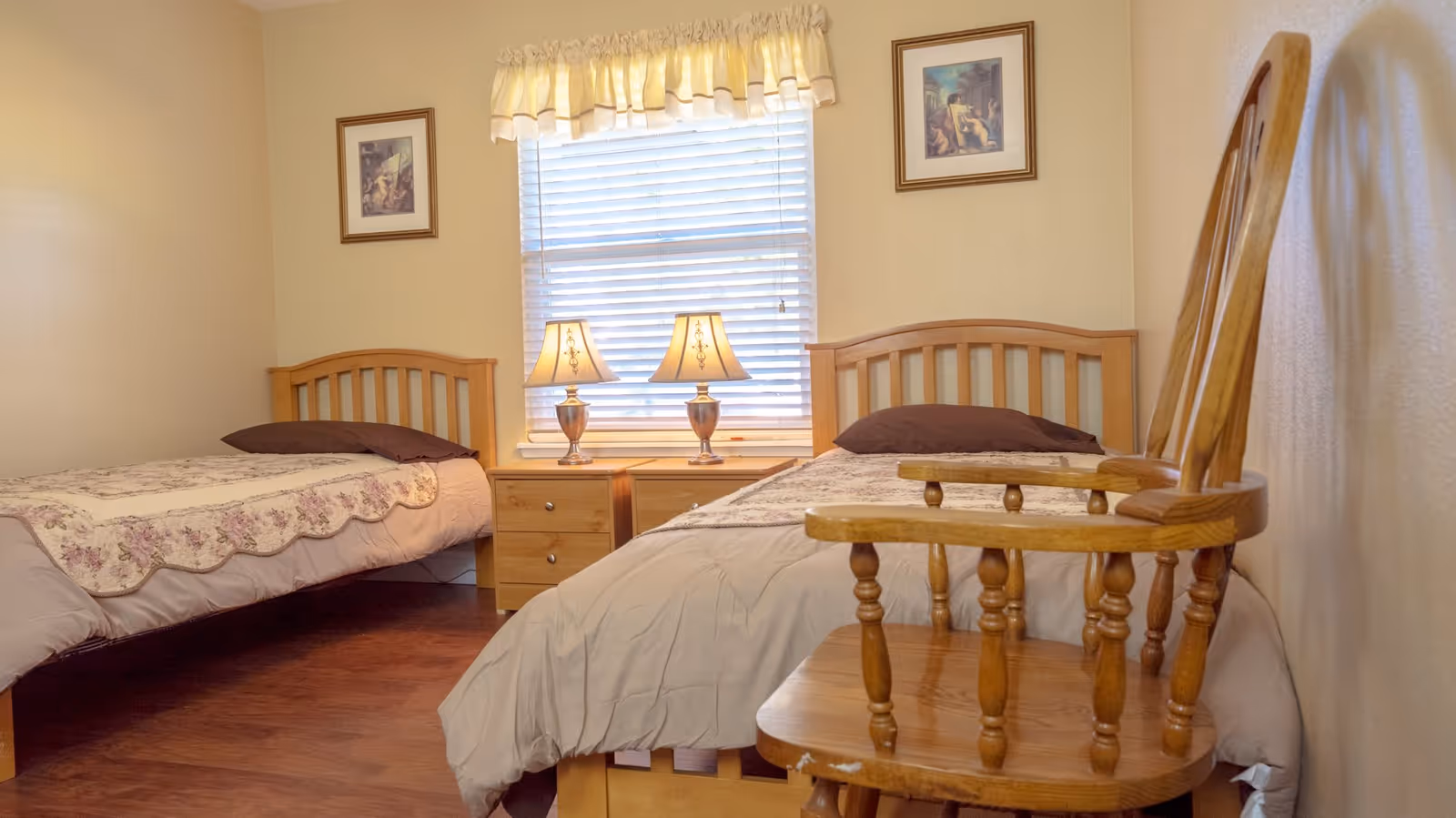 A cozy bedroom with two single wooden beds, each with a floral quilt and a dark pillow. Between the beds is a wooden nightstand with two matching lamps. A window with white blinds and a yellow valance is centered on the wall behind the nightstand. Two framed pictures hang on the wall above each bed. A wooden chair is positioned near the bed on the right side of the image.