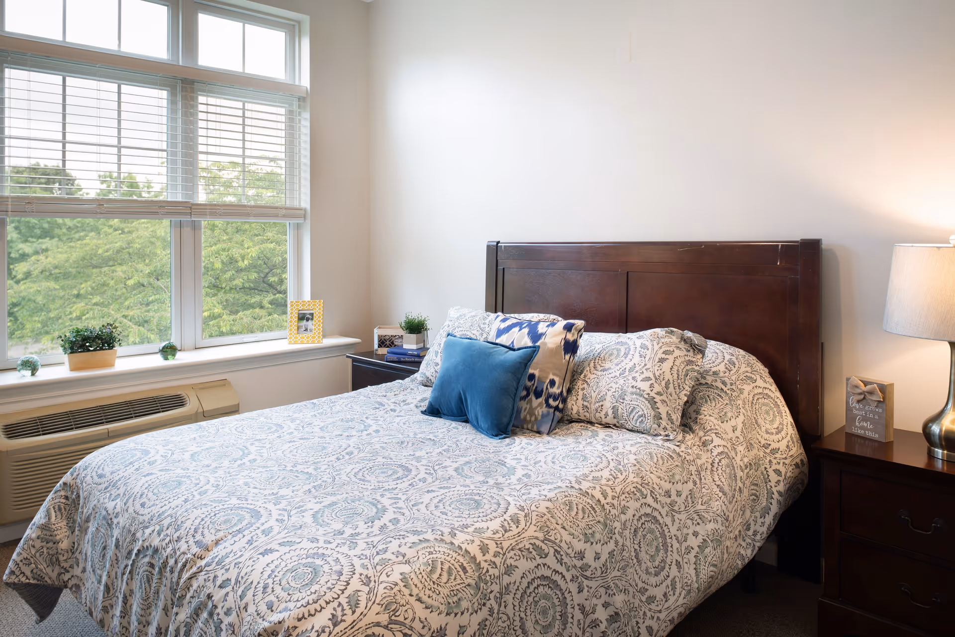 A bright bedroom with a large window showing green trees outside. The room features a bed with a patterned bedspread and multiple pillows, a dark wooden headboard, and matching nightstands. One nightstand has a lamp and a decorative sign, while the other has a small plant and a framed photo.