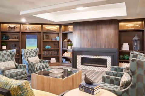 A cozy senior living facility lounge area with patterned armchairs arranged around a wooden coffee table. The room features built-in wooden bookshelves filled with books and decorative items, and a modern fireplace with a tiled surround. The ceiling has recessed lighting and a tray design.