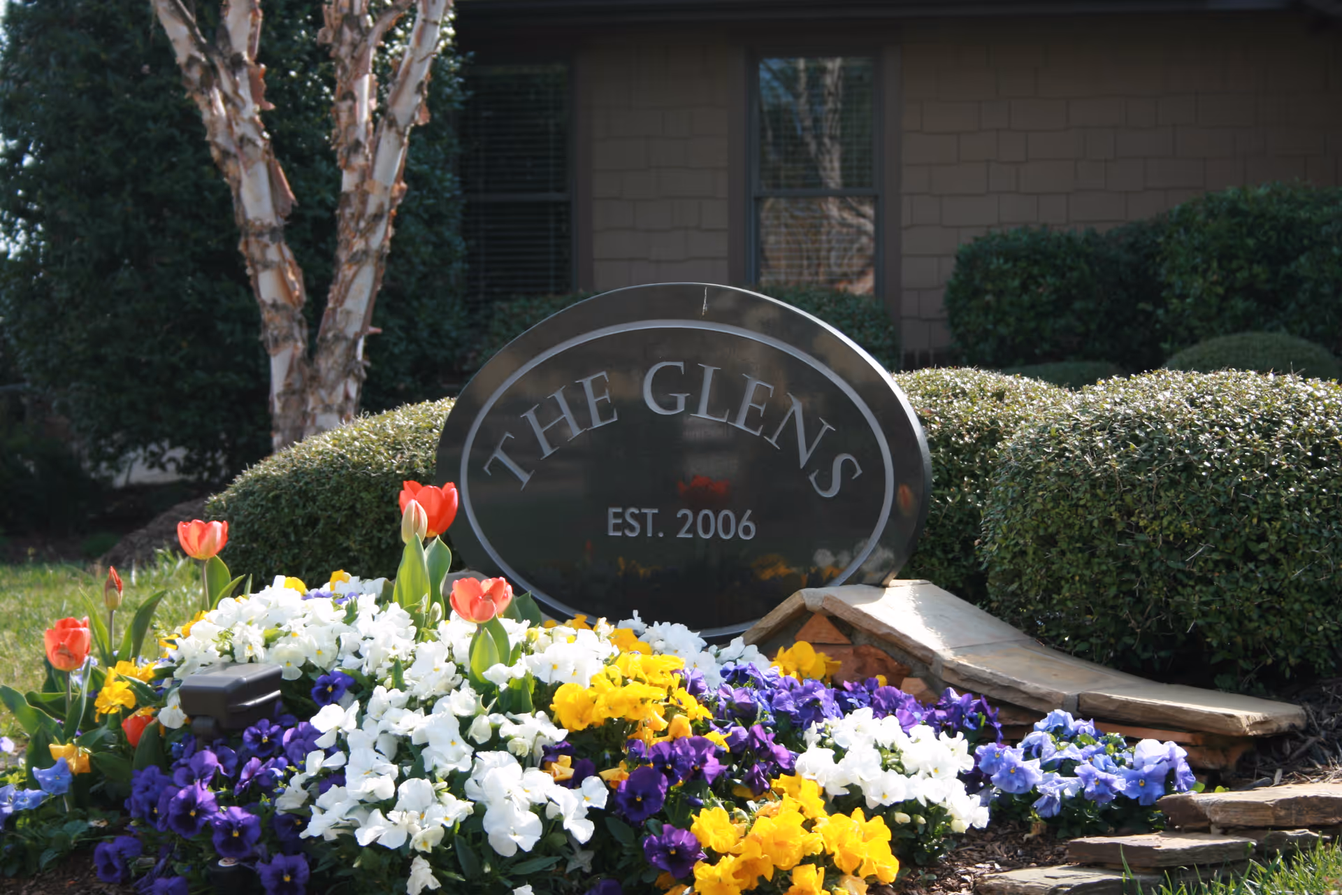 Stone entrance sign reading "The Glens EST. 2006" surrounded by colorful flowers and trimmed shrubs in front of a building.