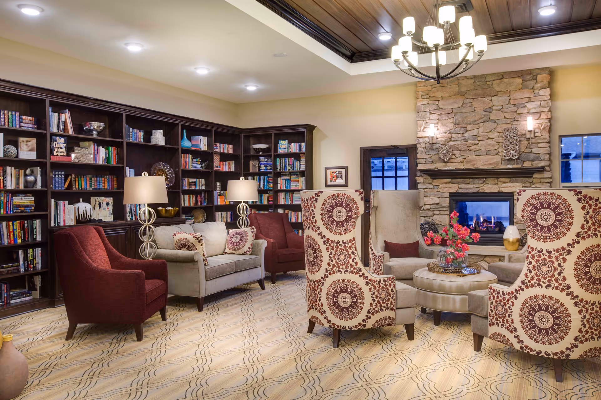 A cozy living room/library with upholstered chairs and sofas, built-in bookshelves, and a stone fireplace.