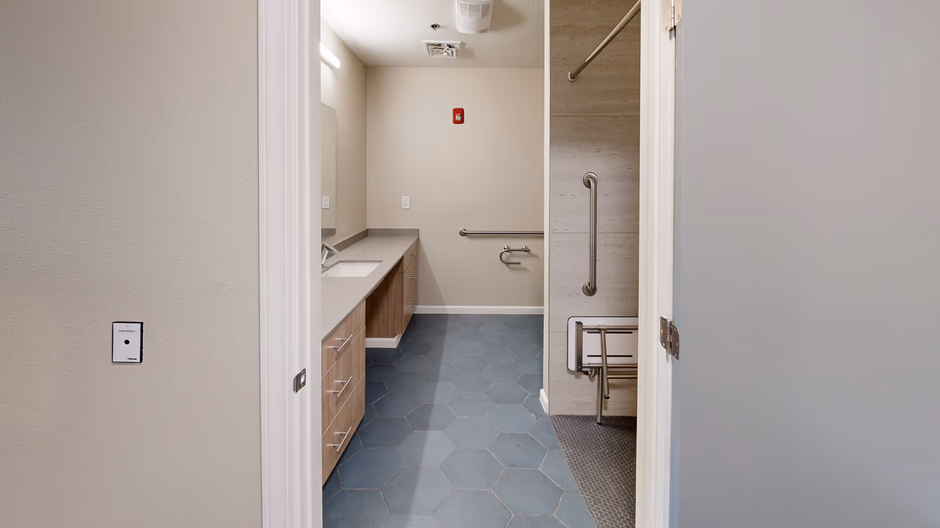 Accessible bathroom with a long vanity and sink on the left, hexagonal tile floor, and a grab-bar-equipped shower with a fold-down seat on the right.
