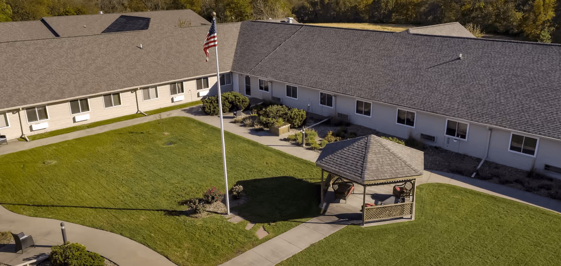 Aerial view of a senior living facility courtyard with a green lawn, a flagpole with an American flag, a gazebo with seating, and a surrounding building with multiple windows.