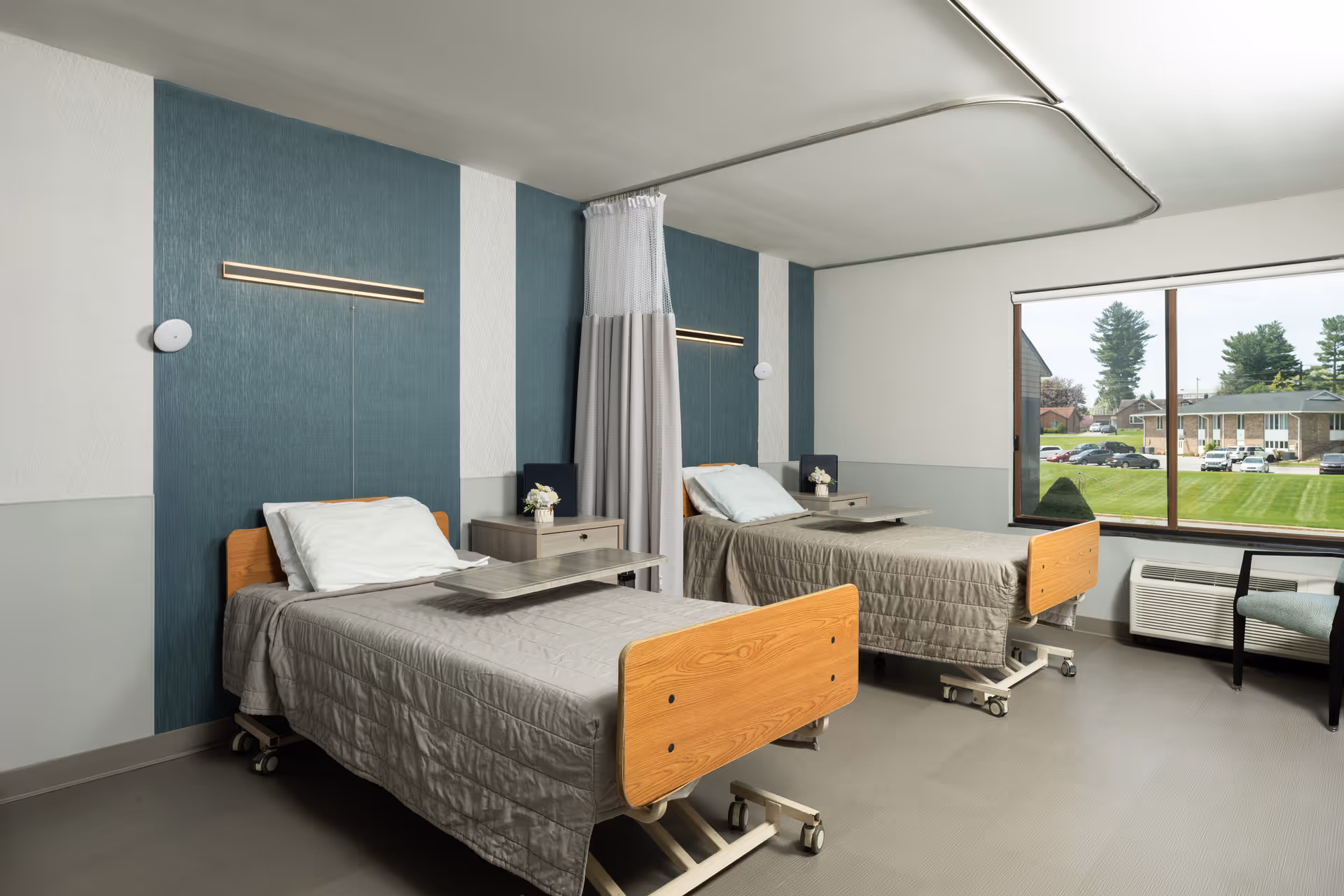 A bright and clean hospital-style room with two adjustable beds, each with gray bedding and white pillows. A curtain divider hangs between the beds. Each bed has a small bedside table with a flower vase. A large window shows a view of a green lawn and nearby buildings. The room has modern lighting and neutral-colored walls.