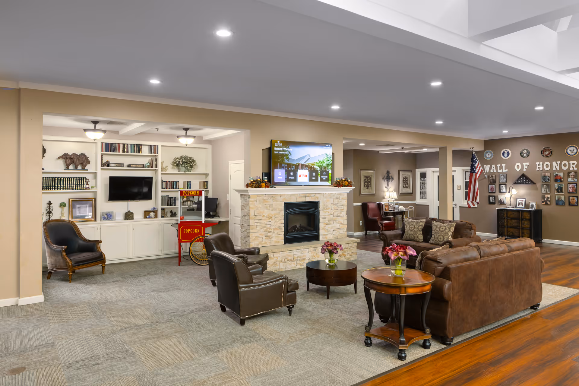 A spacious senior living facility common area with comfortable leather sofas and armchairs arranged around a stone fireplace with a TV mounted above it. There is a popcorn machine near built-in white shelves filled with books and decorative items. The wall in the background features a 'Wall of Honor' with framed photos and an American flag. The room has a mix of carpet and wood flooring, recessed lighting, and a cozy, welcoming atmosphere.