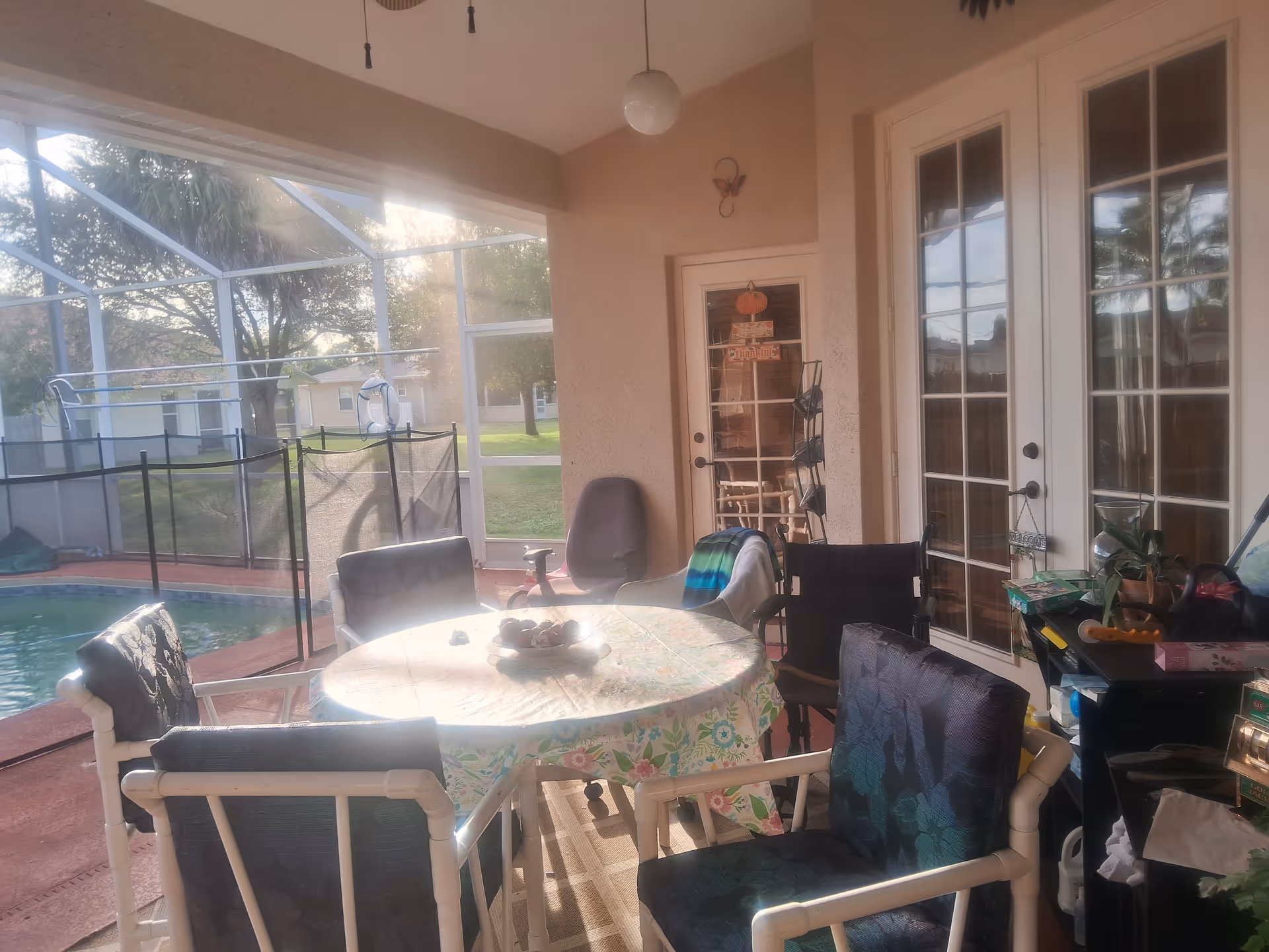 A covered patio area with a round table covered by a floral tablecloth surrounded by six chairs. The patio overlooks a fenced swimming pool and a grassy yard with trees. There are glass doors leading inside the building and various items placed on a side table near the doors.