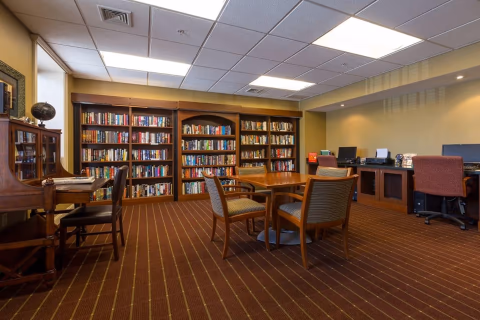 A cozy library or reading room with wooden bookshelves filled with books along the back wall, a round wooden table with four chairs in the center, a wooden desk with a chair on the left, and a workspace with computers and office chairs on the right. The room has a carpet with a striped pattern and a drop ceiling with fluorescent lighting.
