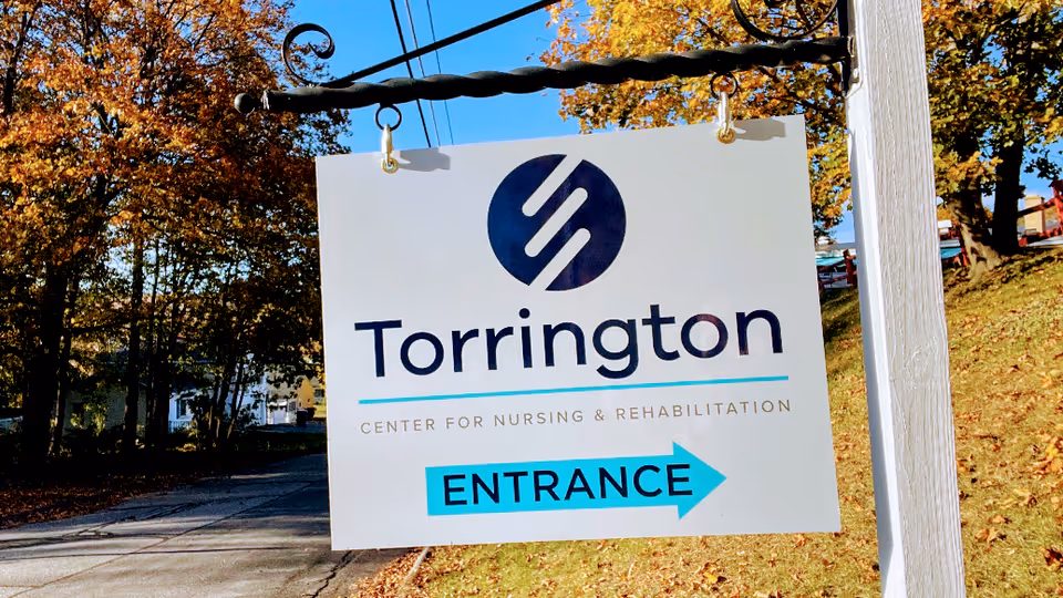 A white hanging sign with the logo and text 'Torrington Center for Nursing & Rehabilitation' and a blue arrow pointing right labeled 'ENTRANCE', set against a backdrop of trees with autumn foliage and a clear blue sky.