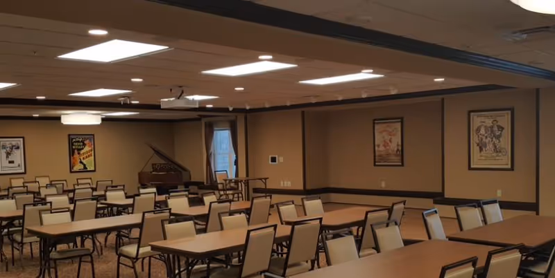 A large room with multiple rectangular tables and beige chairs arranged in rows, beige walls with framed posters, a grand piano in the corner, and a ceiling with recessed lighting and a projector.