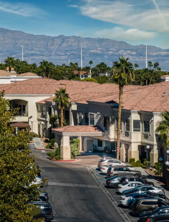 Exterior view of Escalante at the Lakes senior living facility with multiple beige buildings featuring red-tiled roofs, palm trees, parked cars, and mountains in the background under a partly cloudy sky.