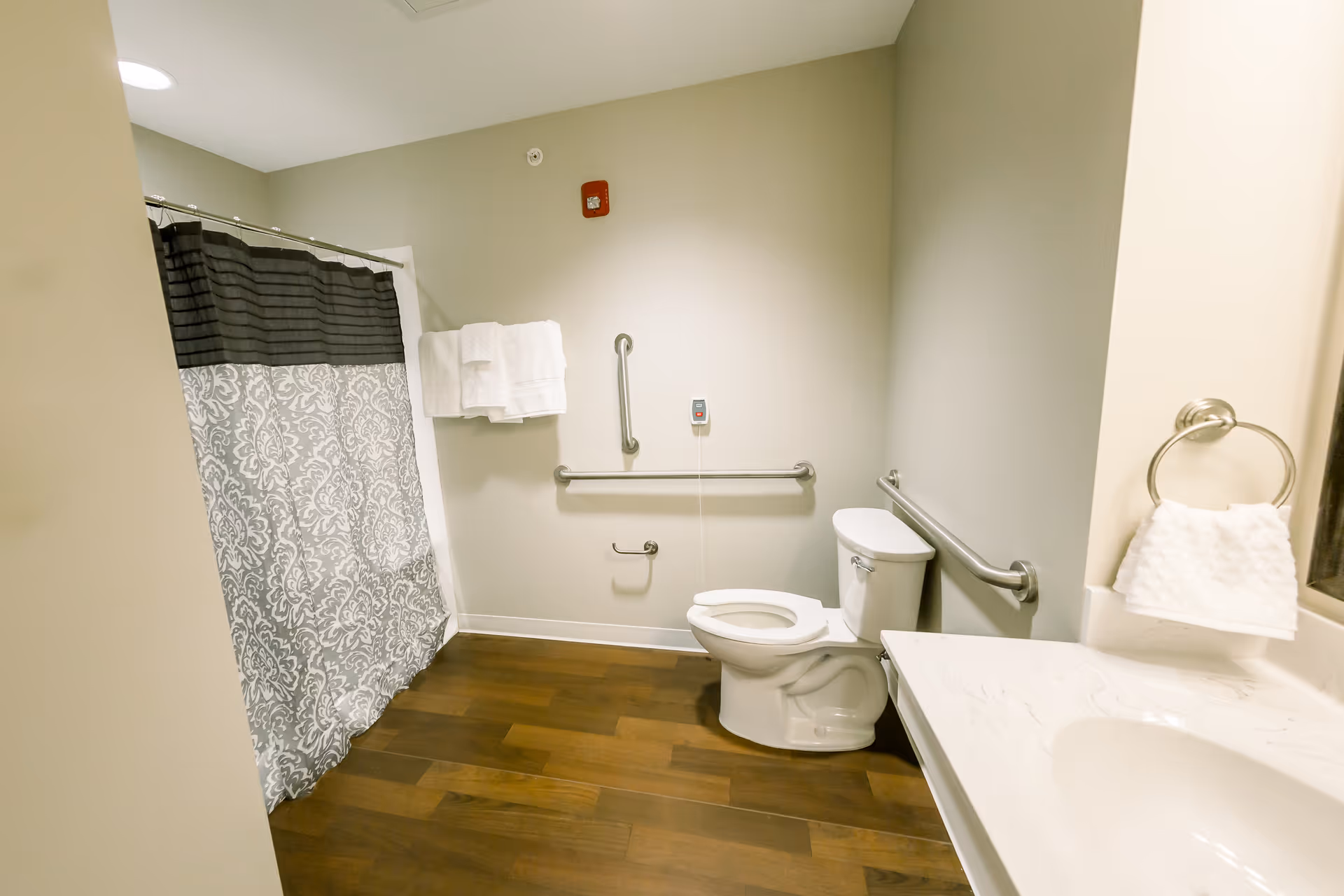 A clean and accessible bathroom with a toilet, grab bars on the walls, a shower with a patterned curtain, a towel rack with white towels, and a sink with a hand towel on a ring holder. The floor is wooden and the walls are painted light gray.