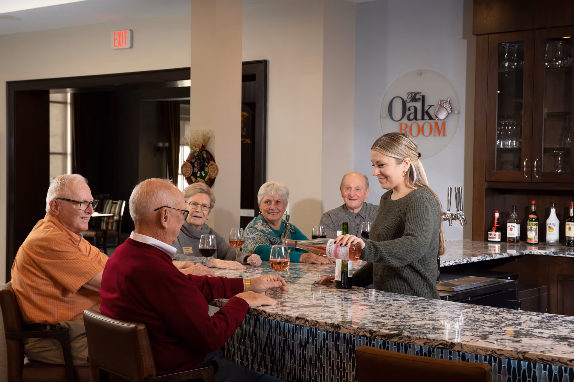 A server pours wine for a group of elderly residents seated at a marble-topped bar in 'The Oak Room' lounge.