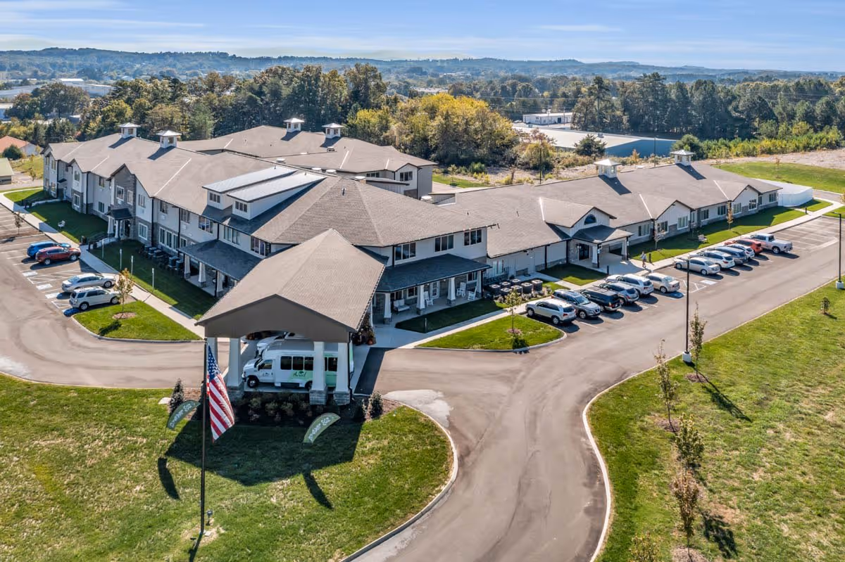 Aerial view of a large senior living facility building with a covered entrance and multiple parking spaces filled with cars. The building is surrounded by green lawns and trees, with a flagpole displaying an American flag near the entrance. The background shows a wooded area and distant hills under a clear sky.