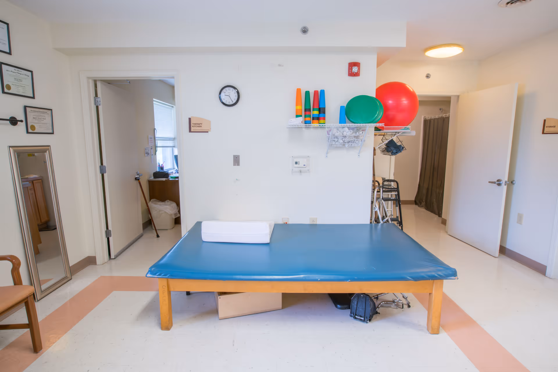 A therapy room in a nursing center featuring a large blue padded therapy table with a white towel on it. The room has white walls, a clock, and shelves holding colorful therapy cones and exercise balls. There are two open doors leading to adjacent rooms, one with a walker visible. Certificates are framed on the wall and a wooden chair is partially visible on the left.