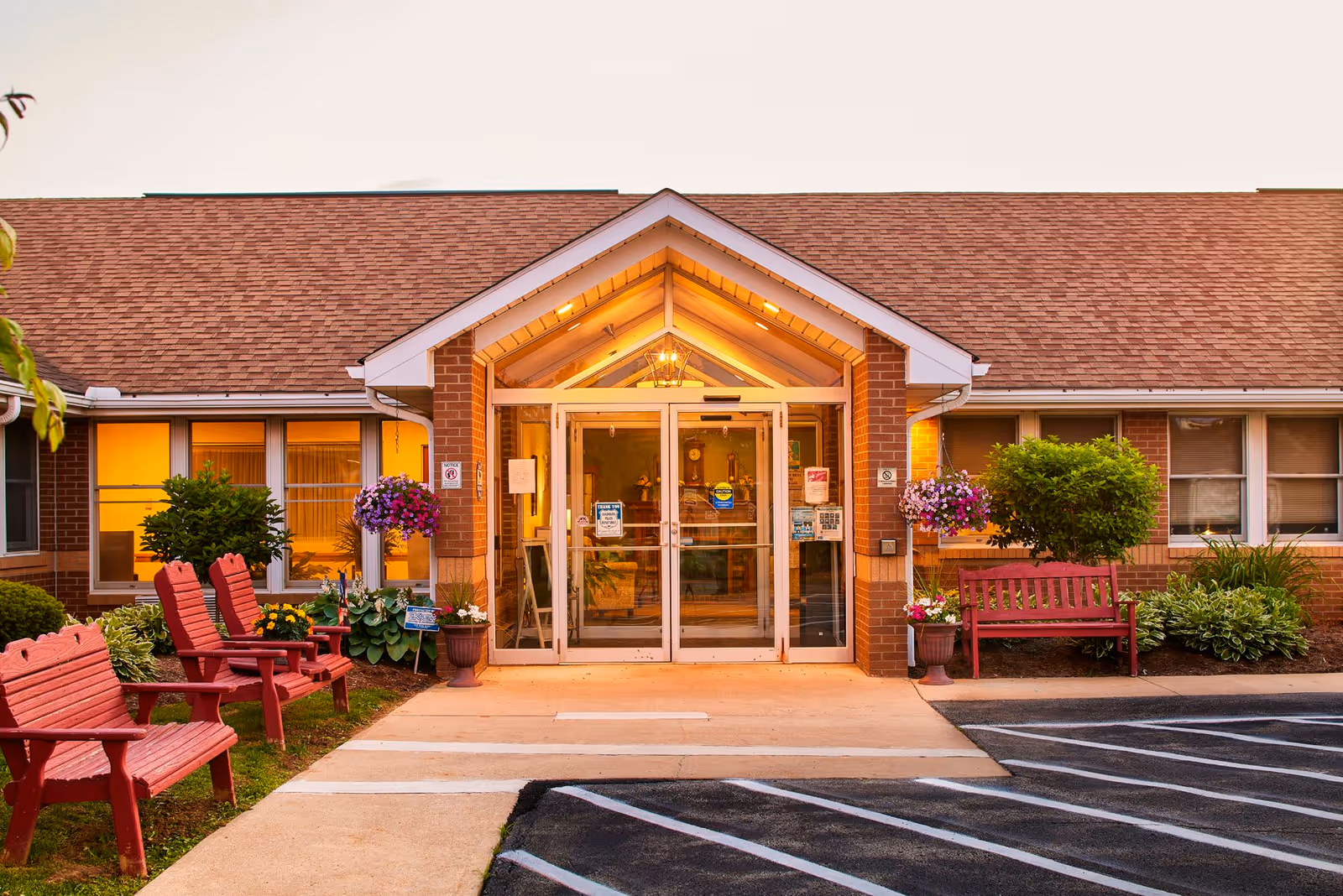 Entrance of Magnolia Place of Saxonburg facility with glass double doors, brick exterior, a peaked roof overhang, red wooden benches and chairs, hanging flower baskets, and landscaped greenery on either side of the walkway.