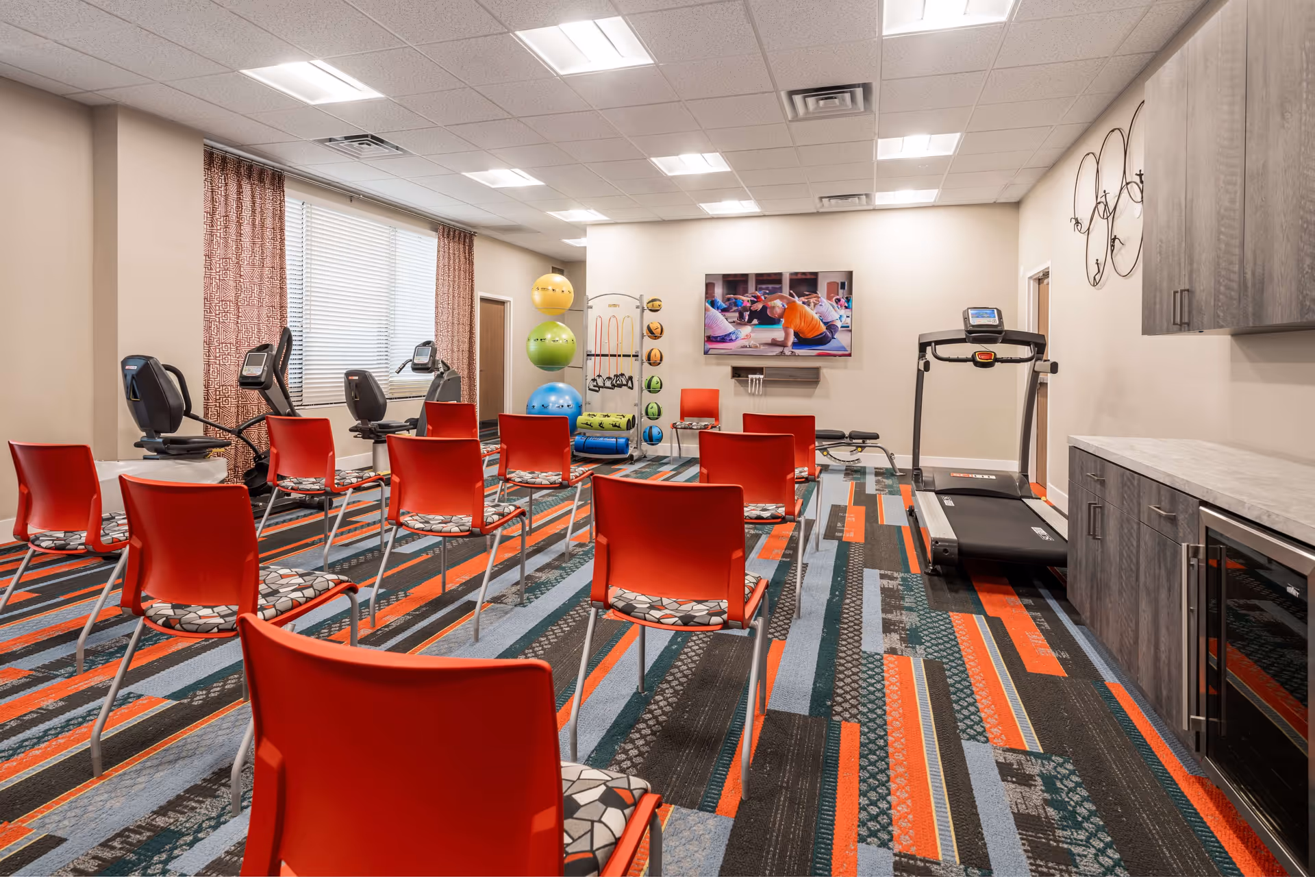 A fitness room with red chairs arranged in rows facing a wall-mounted TV showing a person exercising. The room has exercise equipment including a treadmill, stationary bikes, and various fitness balls and weights. The floor is covered with a colorful patterned carpet, and there are cabinets and a countertop along one wall.