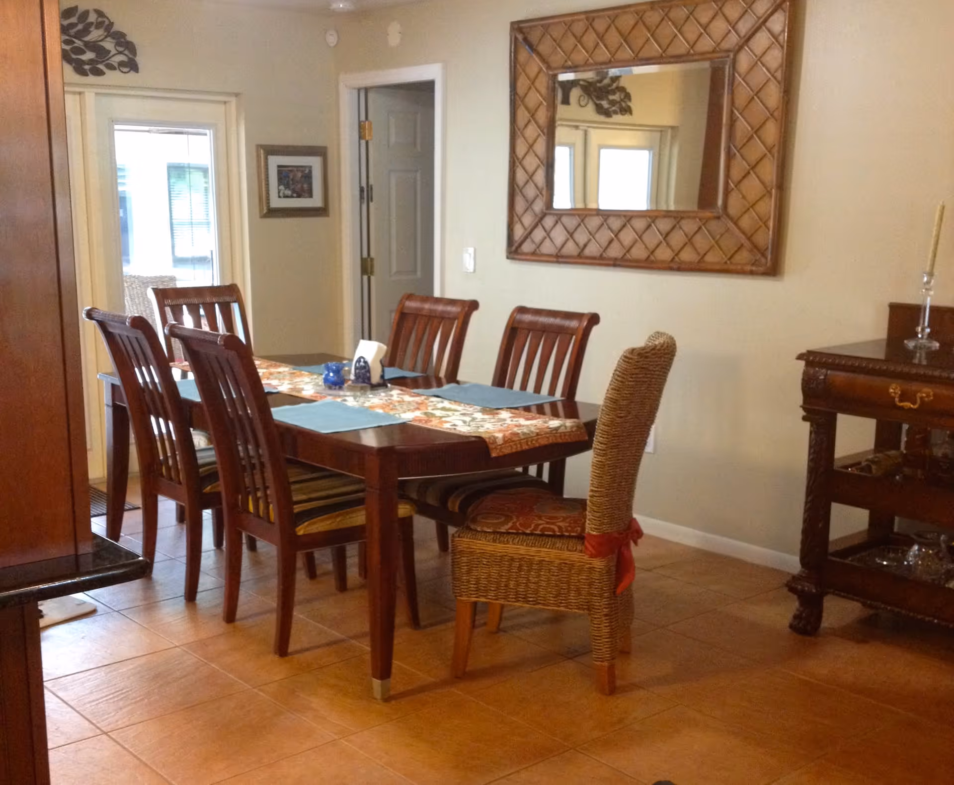 A dining room with a wooden dining table set for six, featuring five wooden chairs and one wicker chair with a red ribbon tied around it. The table has a floral table runner and blue placemats. A large decorative mirror hangs on the wall above a wooden sideboard with a candle holder. There is a glass door and a framed picture on the wall in the background.