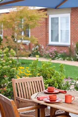 Outdoor patio area with a wooden table and chairs set under a blue umbrella. The table is set with red cups, plates, and napkins. In the background, there is a garden with green shrubs, yellow and pink flowers, and a brick building with white-framed windows.