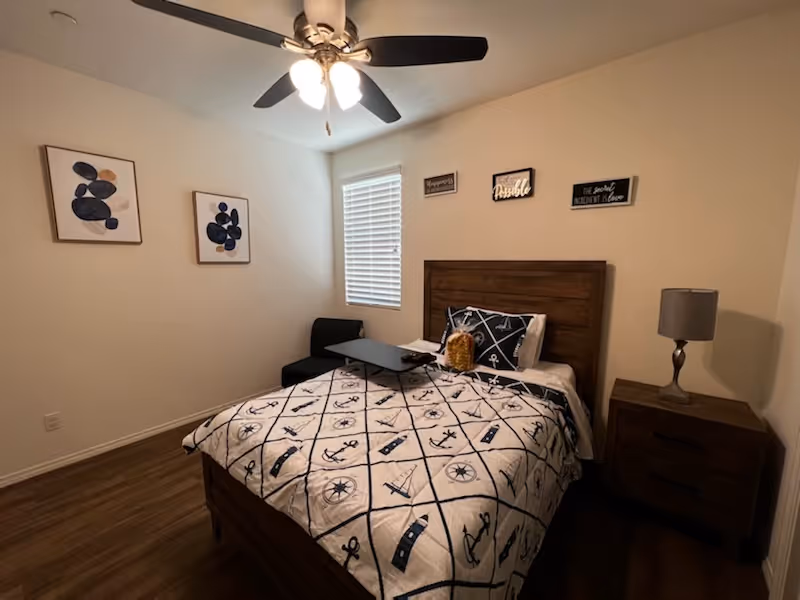A tidy bedroom with a wooden headboard bed dressed in a nautical quilt, nightstand with lamp, ceiling fan, wall art, and a small window.