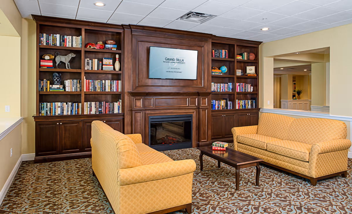 A cozy senior living community lounge with two yellow patterned sofas facing each other across a dark wooden coffee table. Behind the sofas is a large wooden built-in bookshelf filled with books and decorative items, and a fireplace with a mounted flat-screen TV above it displaying the text 'Grand Villa Senior Living Community - St. Petersburg'. The room has a patterned carpet and soft yellow walls.