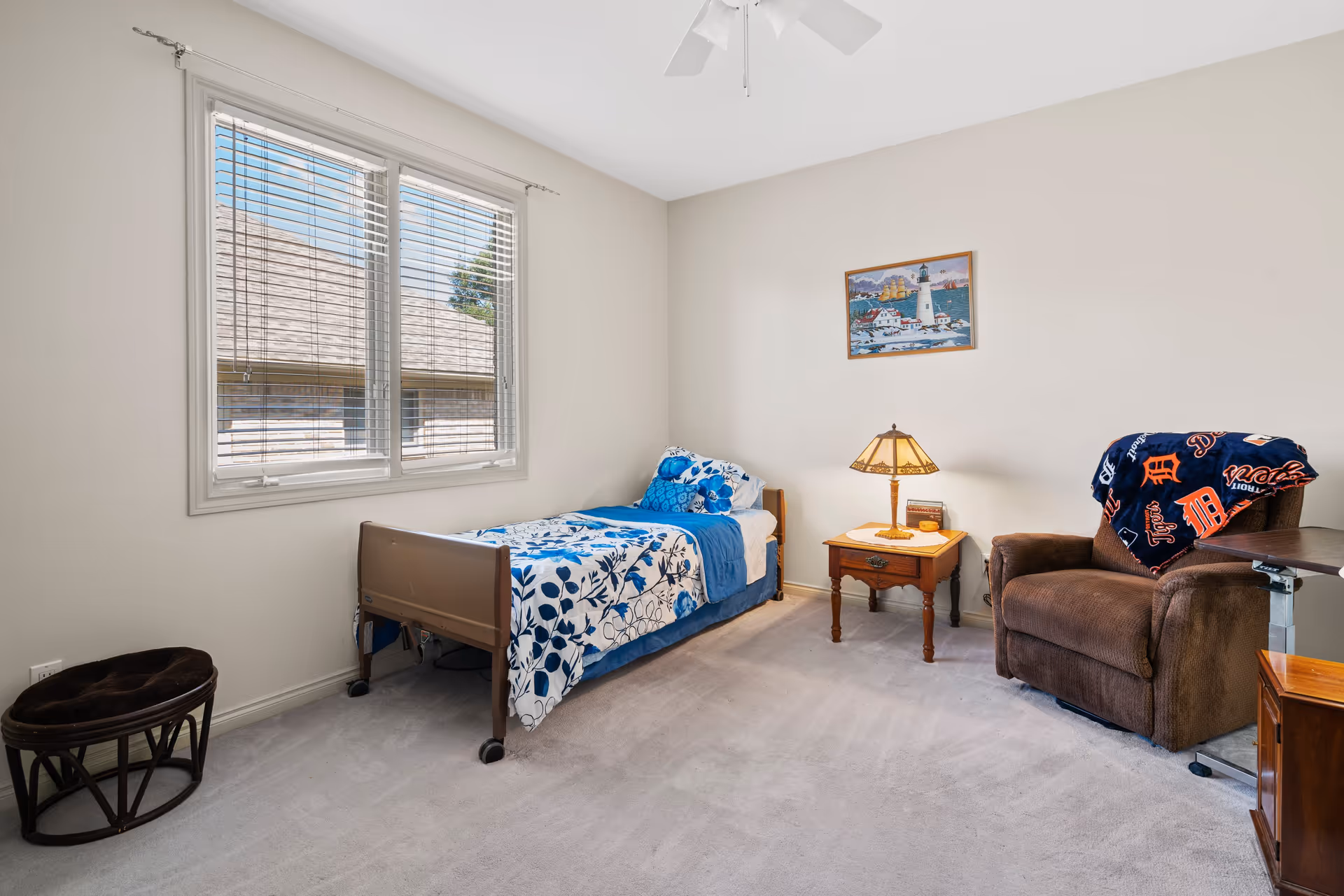 A small bedroom with a single bed covered in blue and white floral bedding. Next to the bed is a wooden nightstand with a decorative lamp and some books. A brown recliner chair with a Detroit Tigers blanket draped over it is positioned to the right of the nightstand. There is a large window with white blinds on the left wall, and a framed picture of a lighthouse hangs on the wall above the nightstand. The room has light-colored walls and carpeted floor.