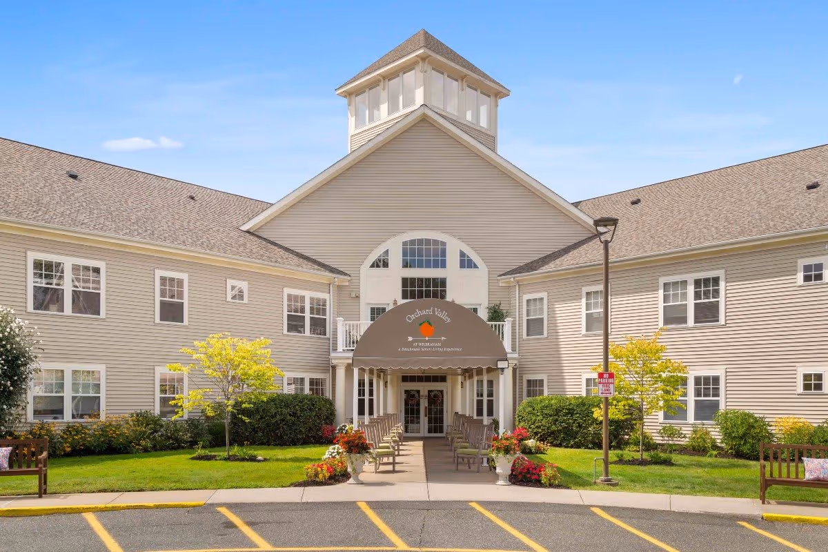Front exterior view of a senior living facility named Orchard Valley with beige siding, multiple windows, a peaked roof with a cupola, a covered entrance with benches on either side, and landscaped greenery including small trees and flower pots.