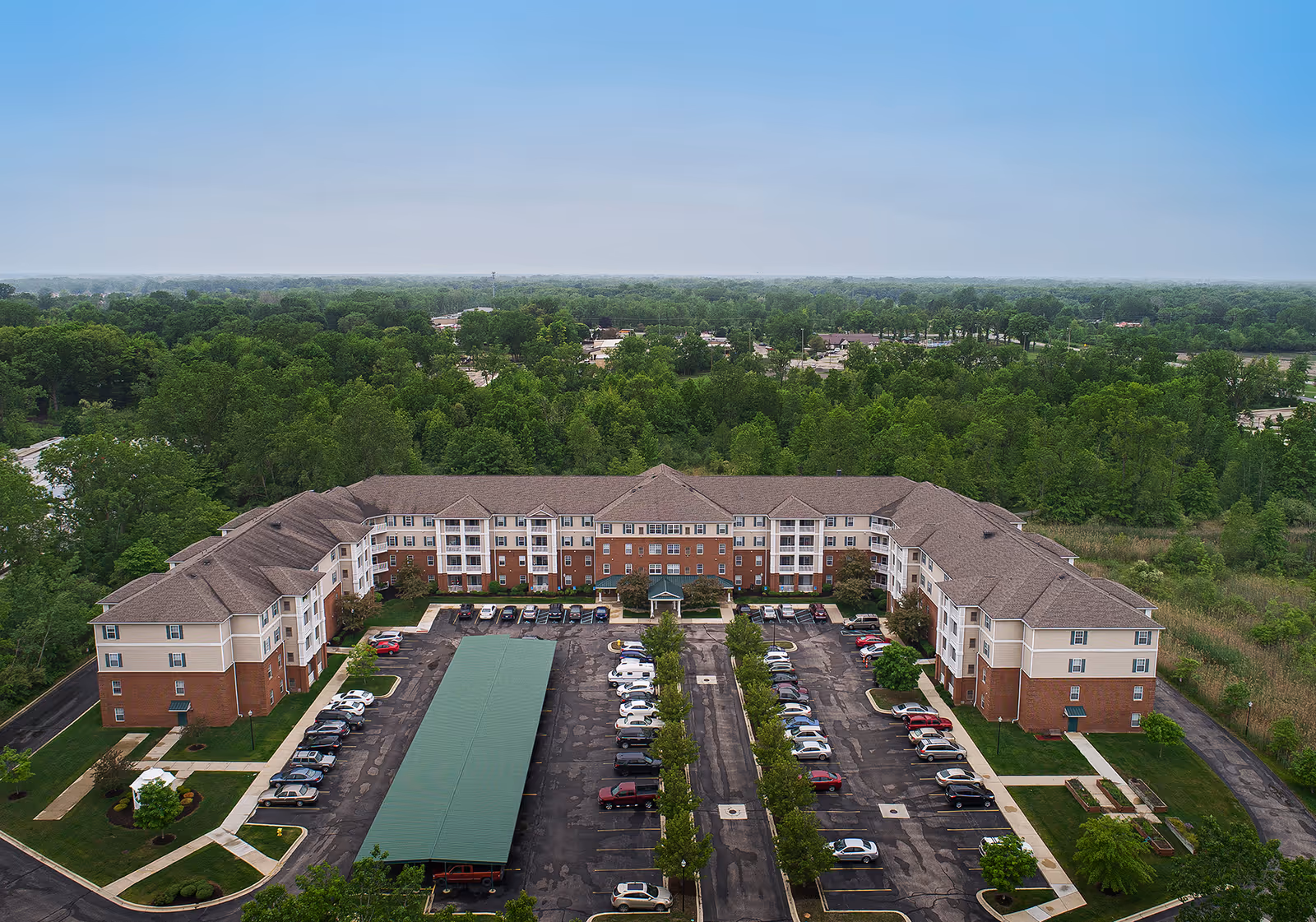 Aerial view of The Meadows of Brownstown senior living facility showing a large U-shaped building with a parking lot in front, surrounded by green trees and vegetation under a clear sky.
