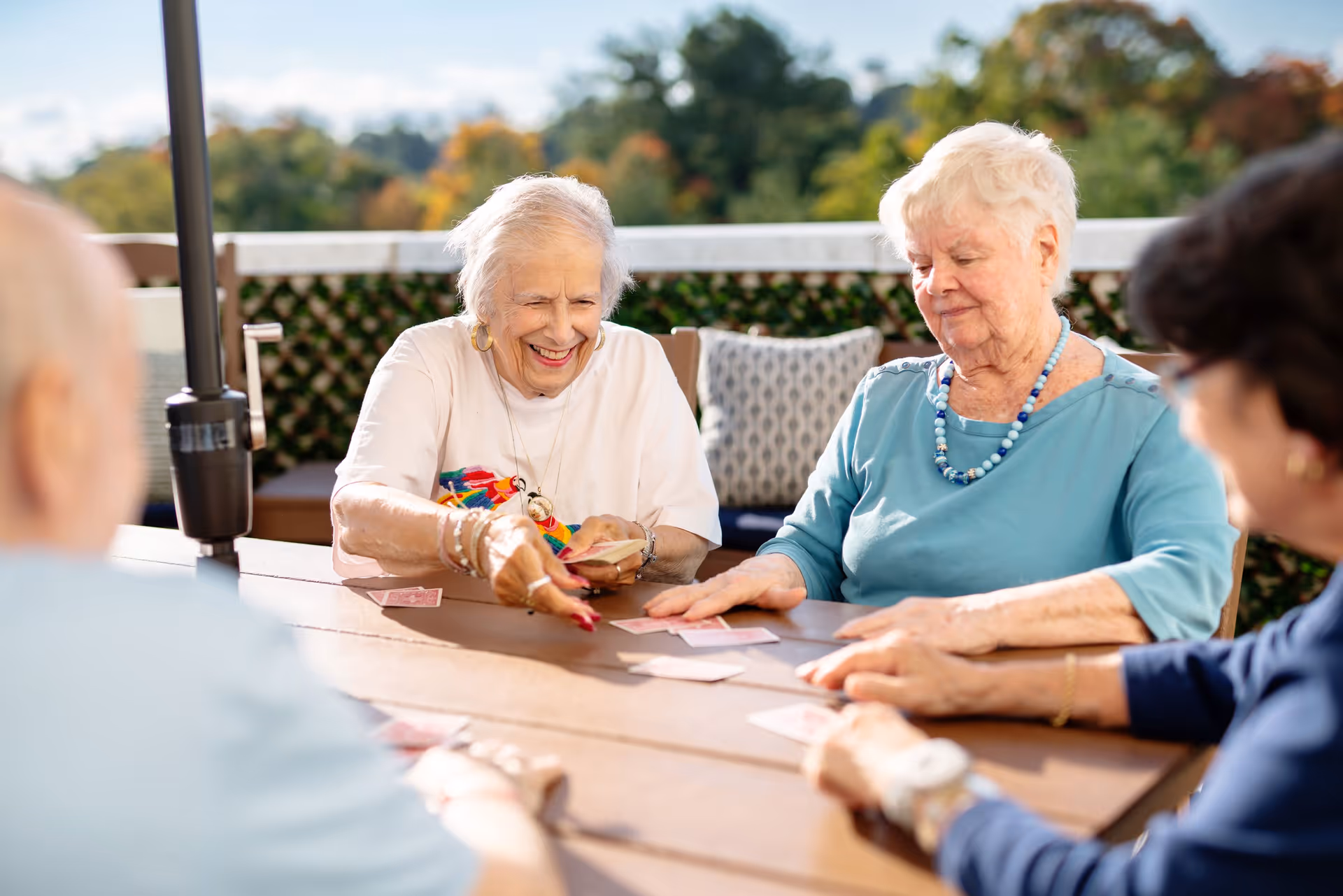 Four elderly women sitting around a wooden table outdoors playing a card game, with greenery and trees in the background under a clear sky.