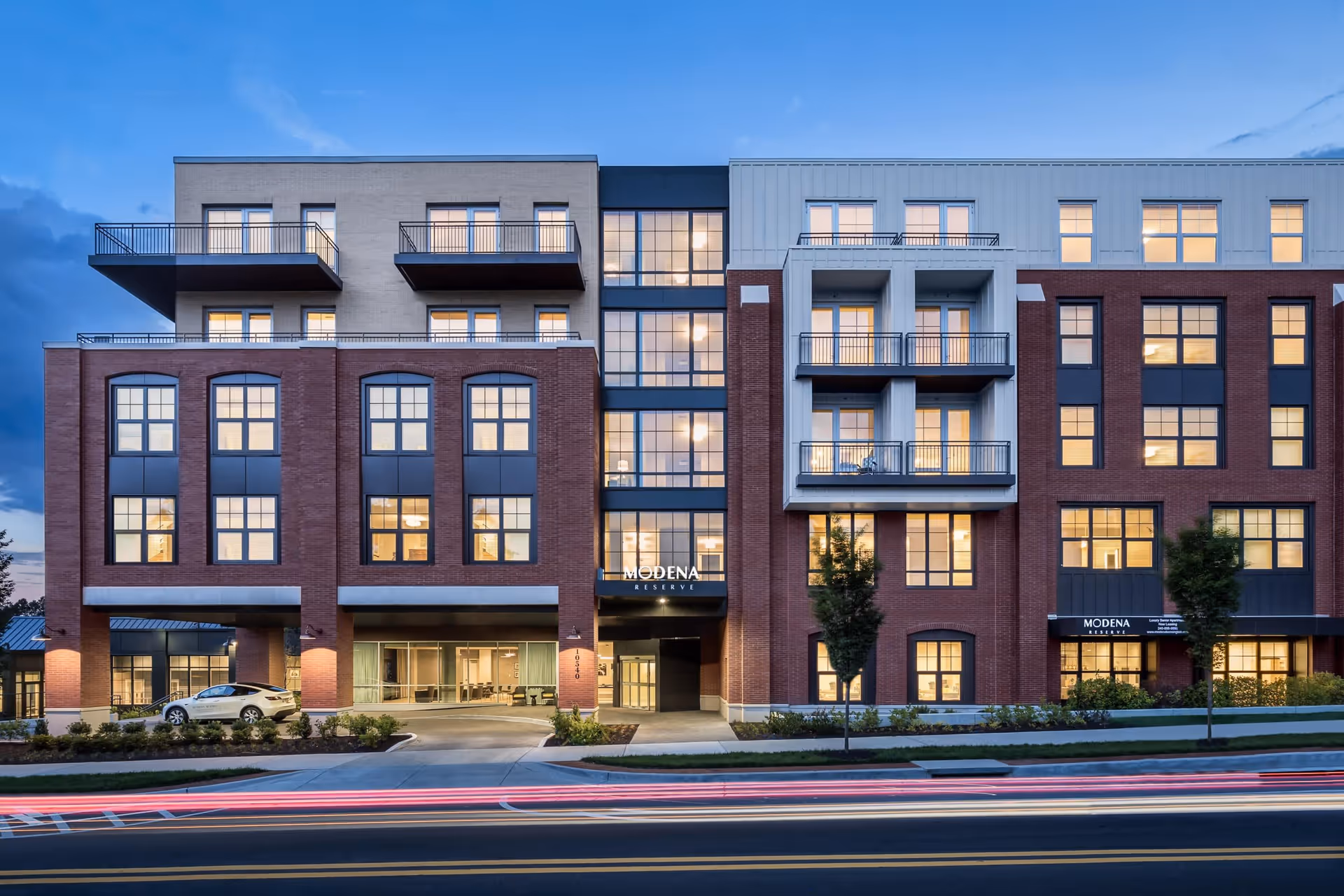 Front facade of a multi-story brick senior living building with balconies and illuminated windows at dusk.